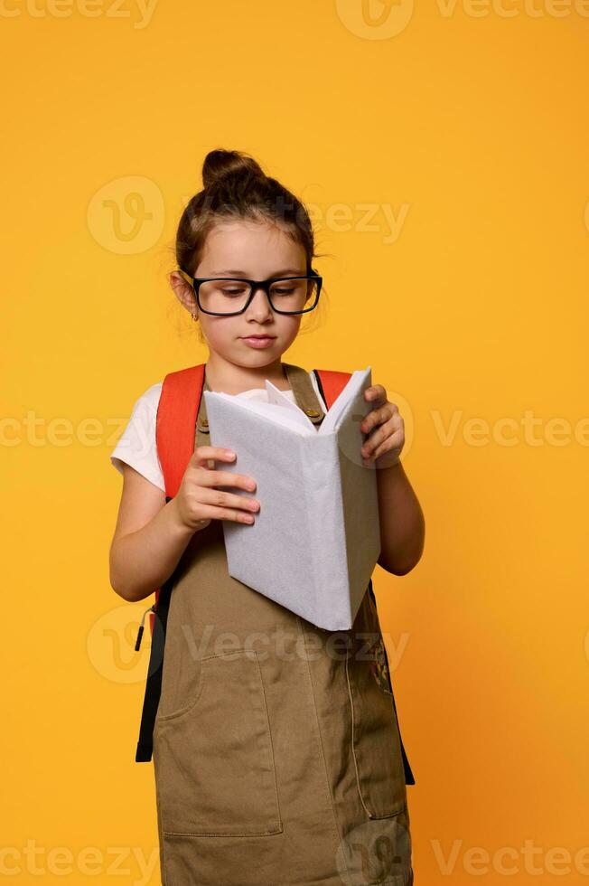 Confident school girl carries school bag, reads book, standing isolated on orange background. Kids and education concept photo