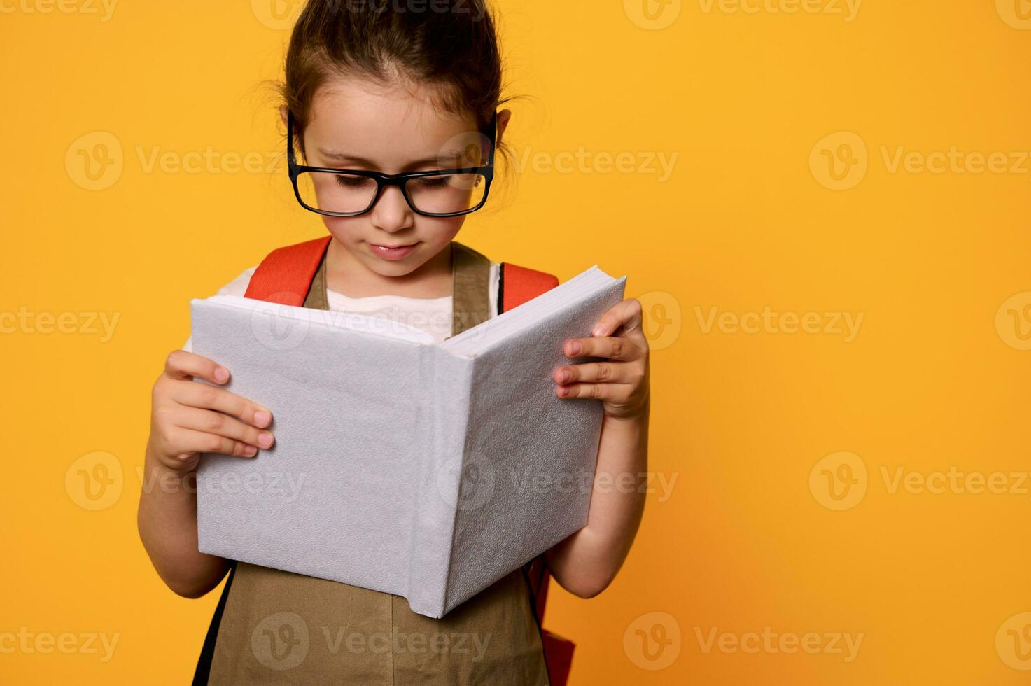 Adorable kid girl in casual wear and glasses, learns reading alphabet, holding mockup book, isolated orange background photo