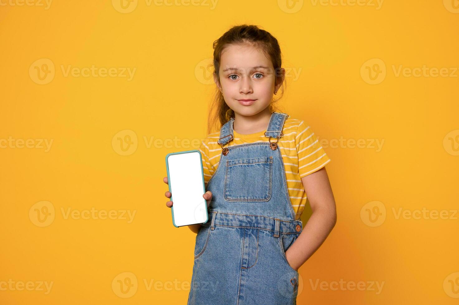 Cute little child girl in casual denim, puts her hand in pockets, showing at camera a white blank mockup digital screen photo