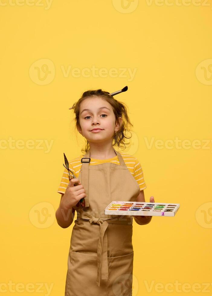 Inspired creative talented kid girl holding watercolor palette, paint brush and palette knife, isolated yellow backddrop photo