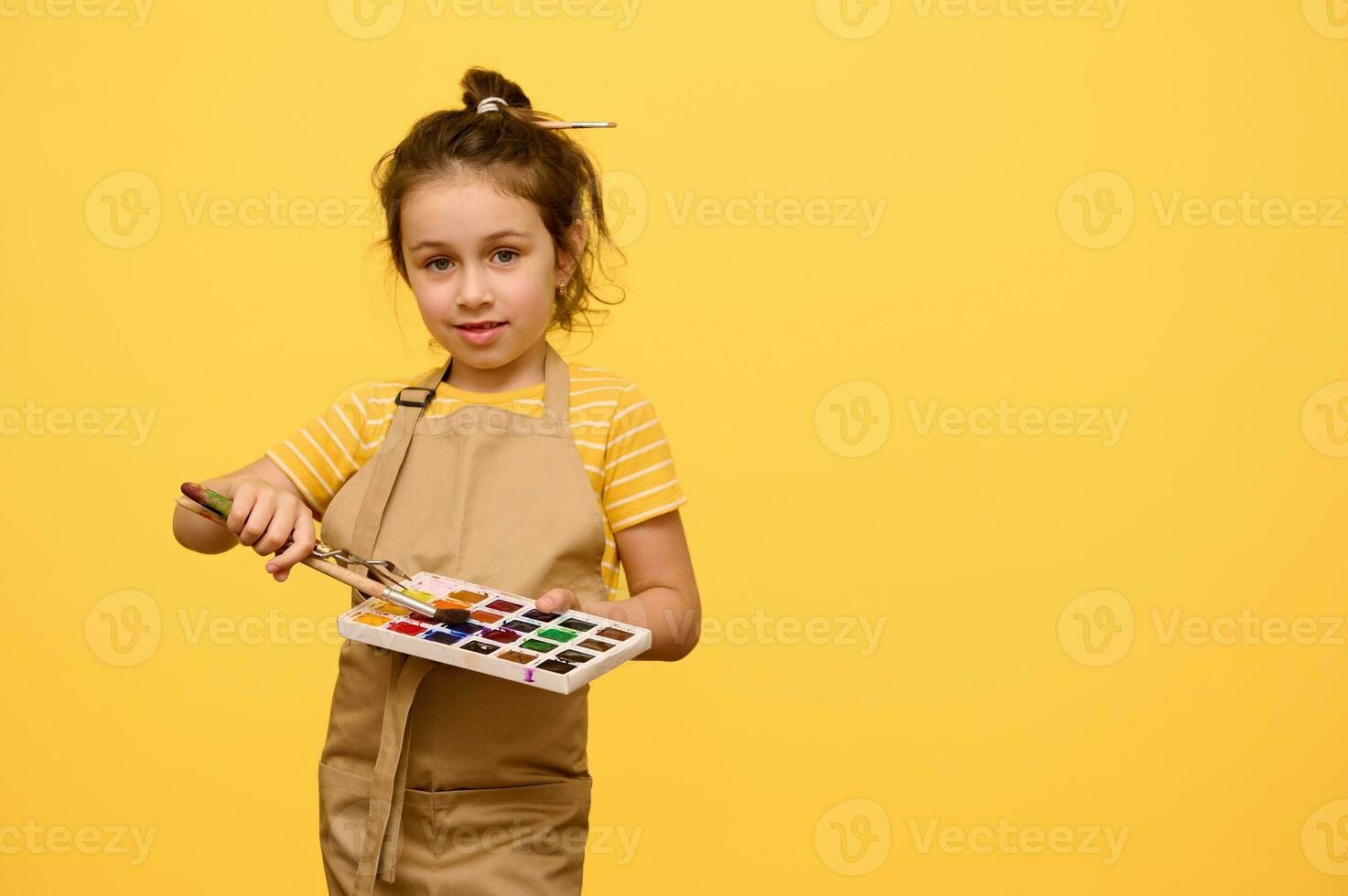 Talented little girl painter artist in apron with paintbrush in hairstyle, holds watercolor palette and painting tools photo