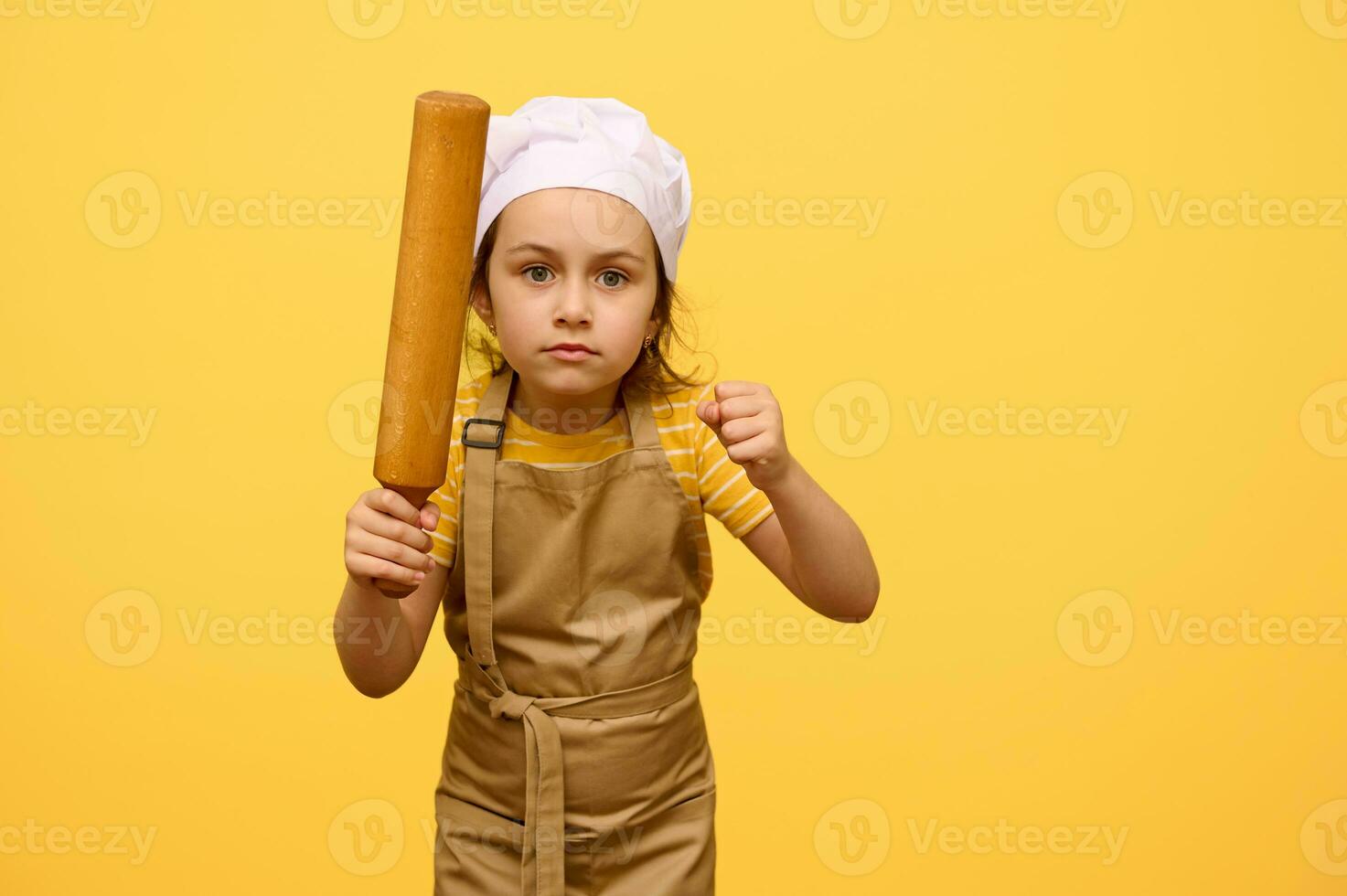 Serious angry little child girl in chef's uniform, holding a wooden rolling pin, clenching fists, looking at camera photo