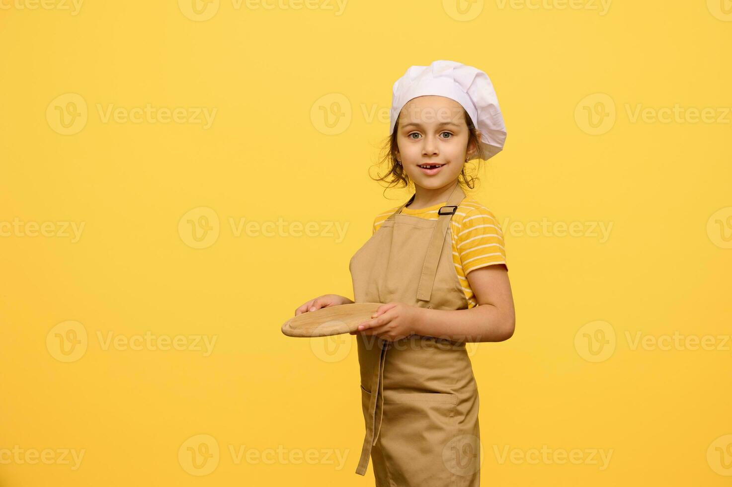Adorable kid schoolgirl dressed as chef confectioner, holds a wooden board, smiles looking at camera, isolated on yellow photo