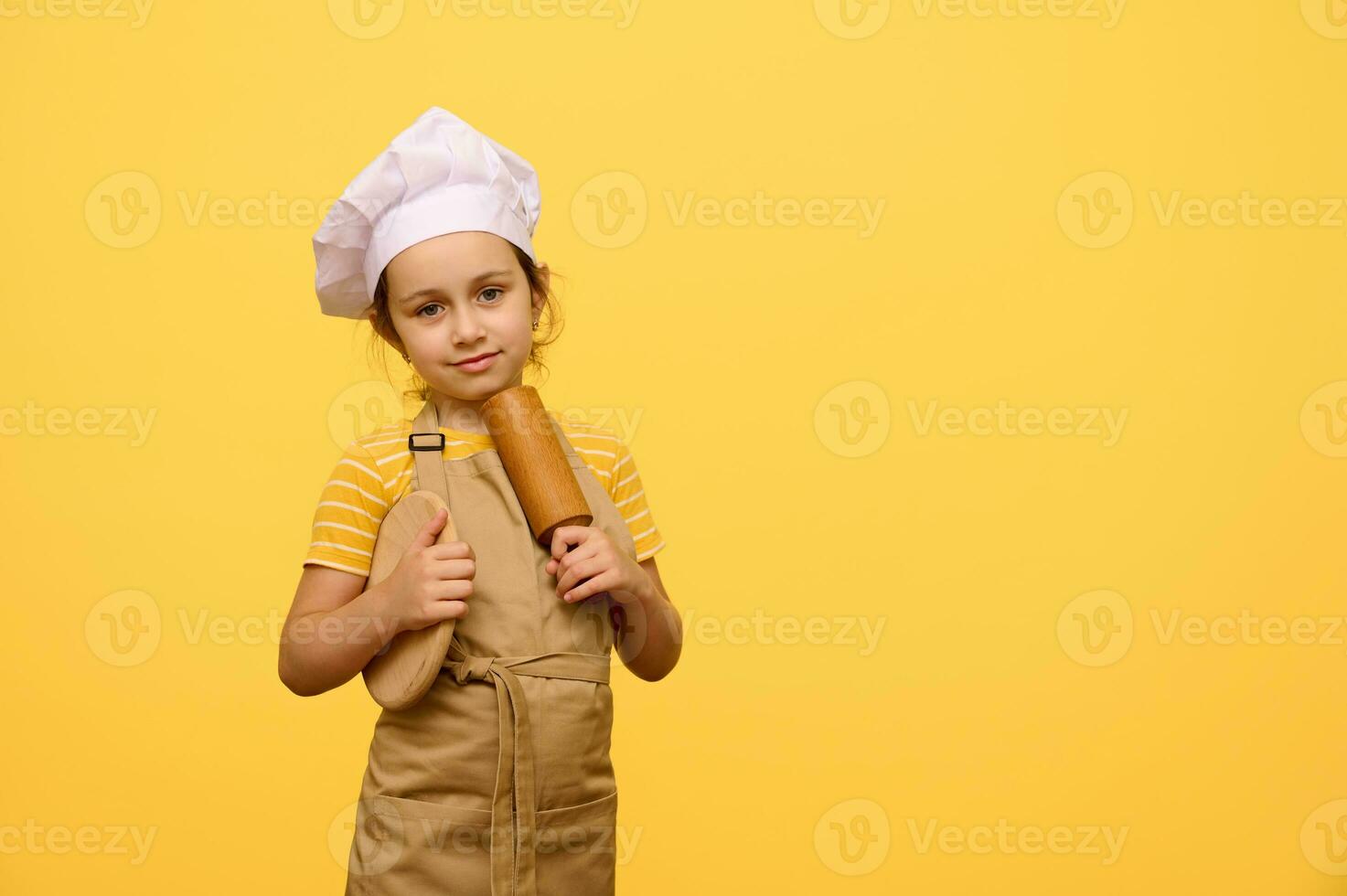 Adorable little baker confectioner girl in beige apron and hat, holding wooden board and rolling pin, smiles at camera photo