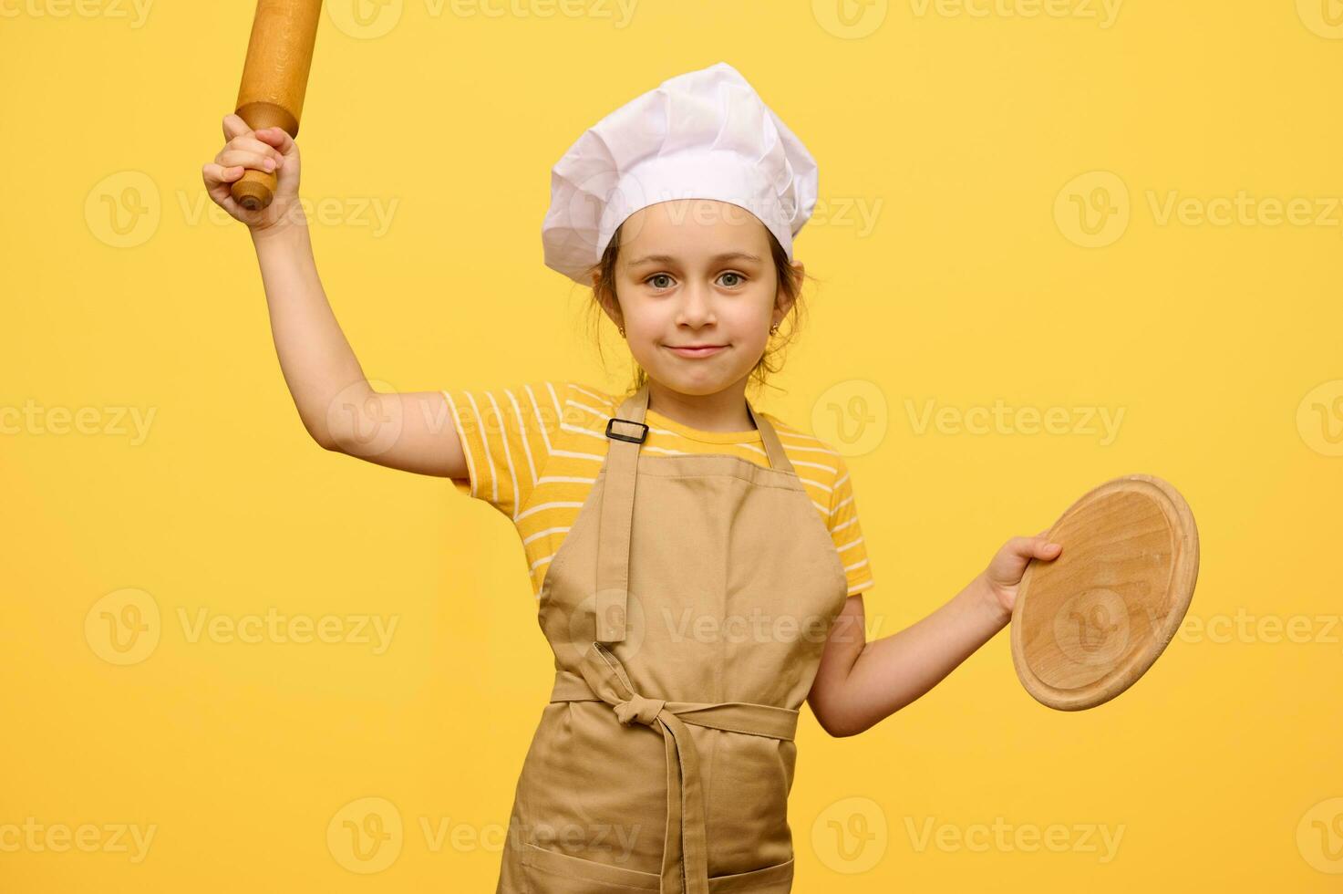 Beautiful child girl in chef hat and apron, smiling at camera, holding rolling pin and wooden board, isolated on yellow photo