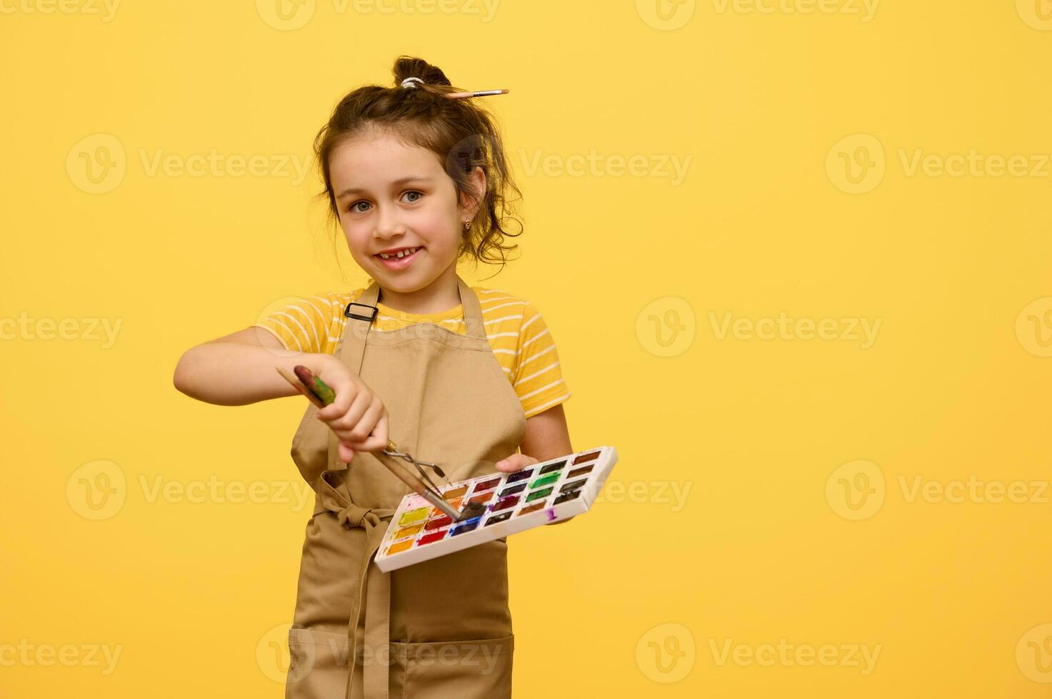 Adorable little girl in beige apron, primary school student dipping paintbrush into watercolor paints, smiles at camera photo