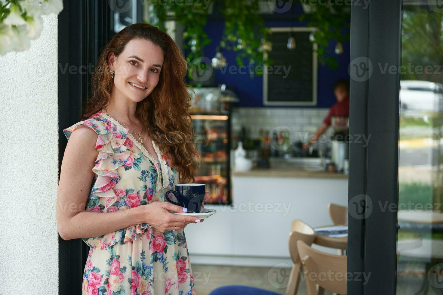 joven sonriente mujer en pie con un taza de café en su manos a el Entrada de su cafetería foto