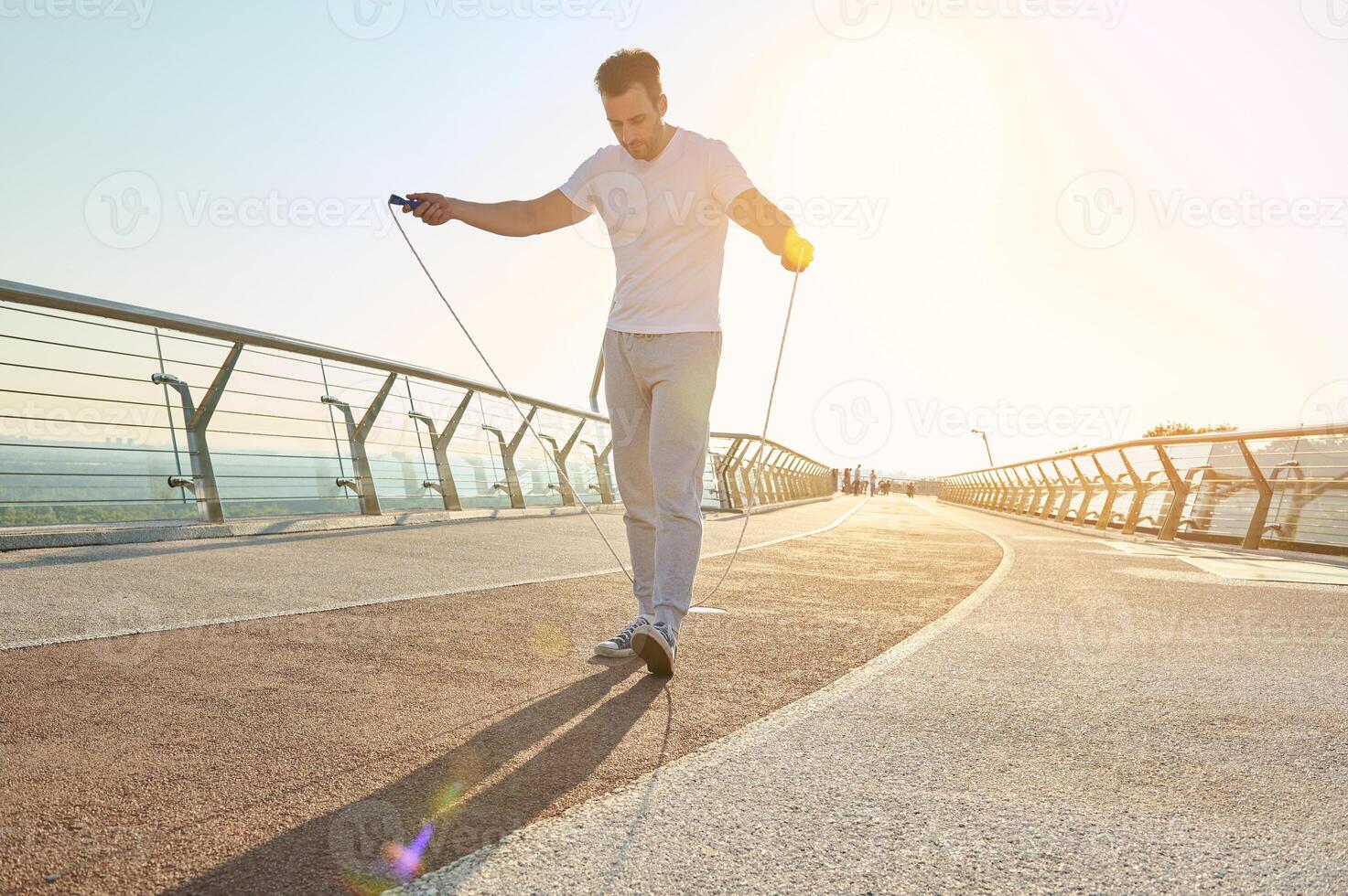 Full length portrait of a muscular build European middle aged man, fit athlete doing jumping exercises, cardio training with skipping rope on the urban city bridge early in the morning on a summer day photo