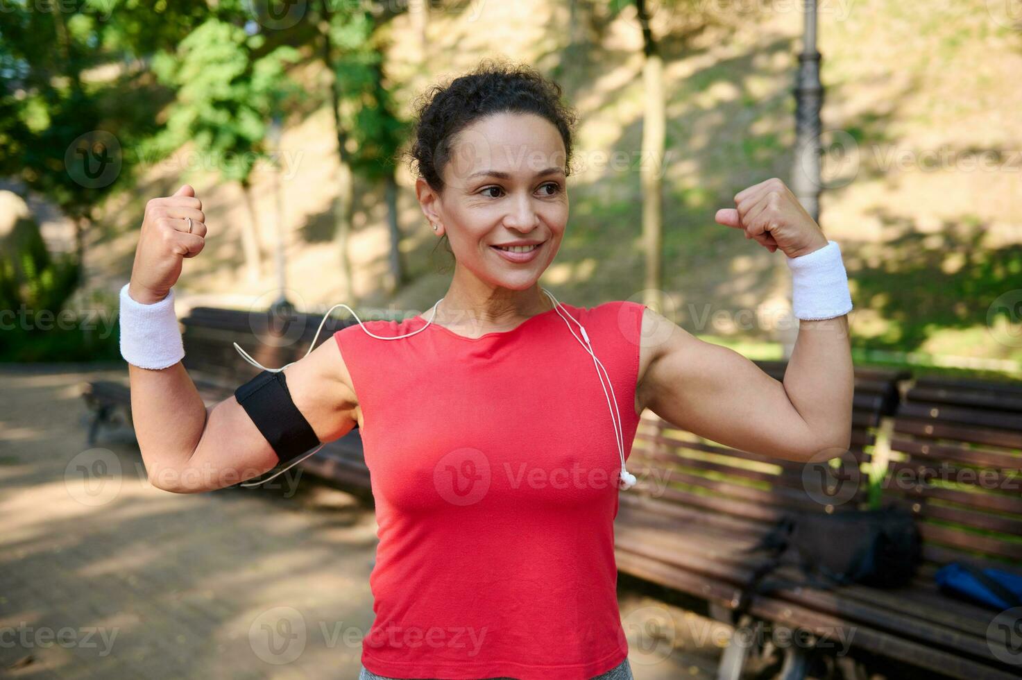 Charming confident active woman, sportswoman clenching fists, tensing arm and biceps muscles, smiling standing on the city park during sports training photo