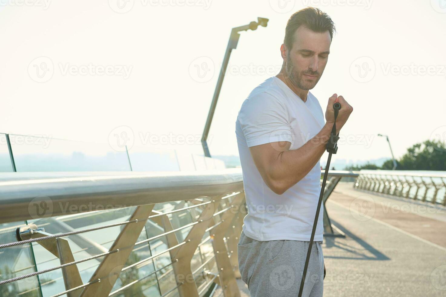 Handsome European young sportsman, athlete during outdoor workout with rubber resistance band. Middle-aged sportsman doing exercises on biceps using an elastic expander on a glass city bridge at dawn photo