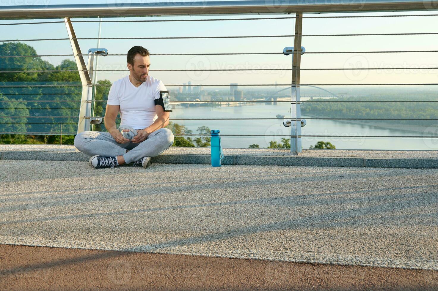 Middle aged Caucasian man in sportswear sitting on a city bridge and checks a mobile application with monitoring heart rate and calories burned during a workout on his phone in a smartphone holder photo