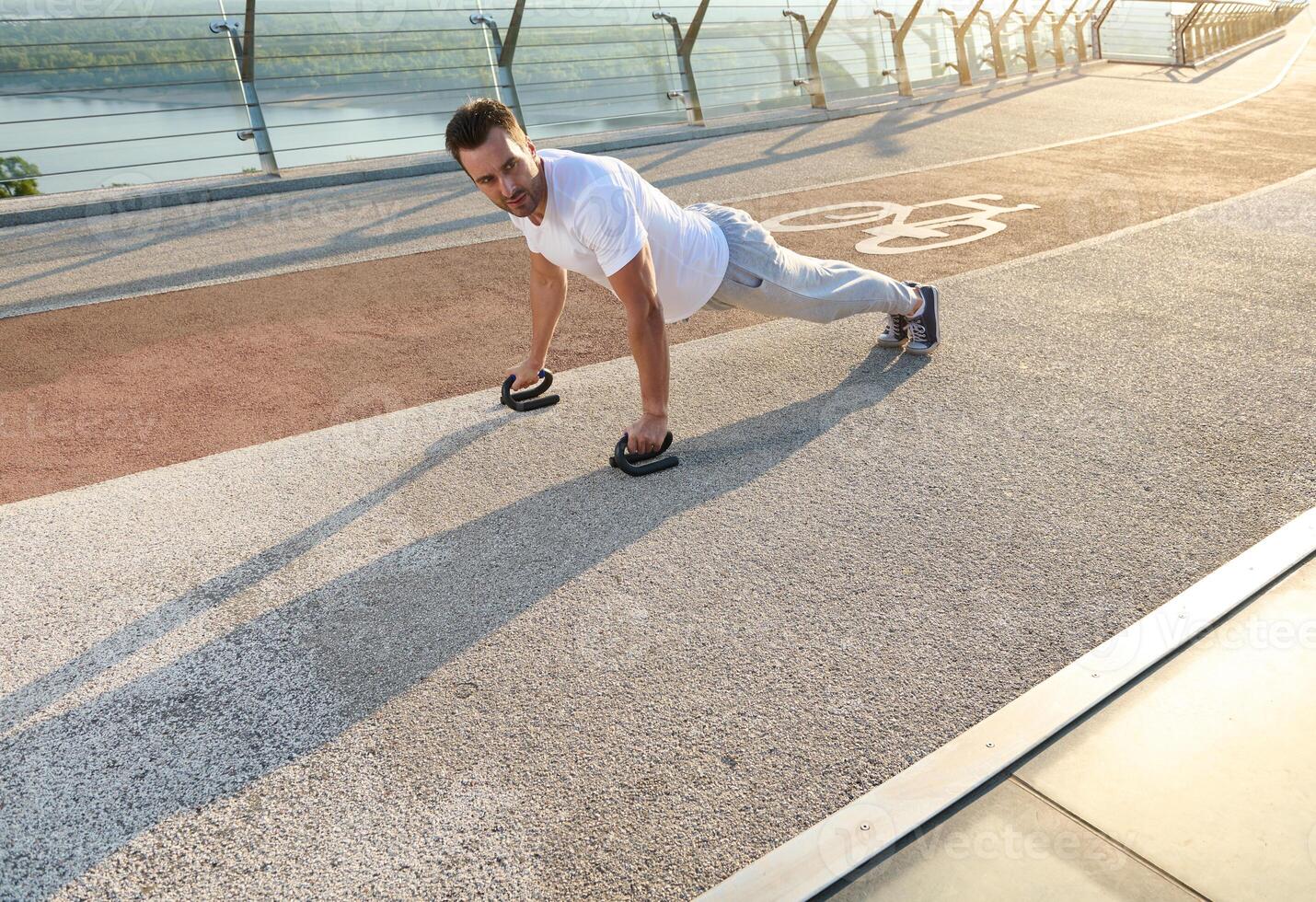 Full length portrait of a handsome middle aged European man, muscular build athlete doing push ups during an outdoor workout on the city bridge early in the morning photo