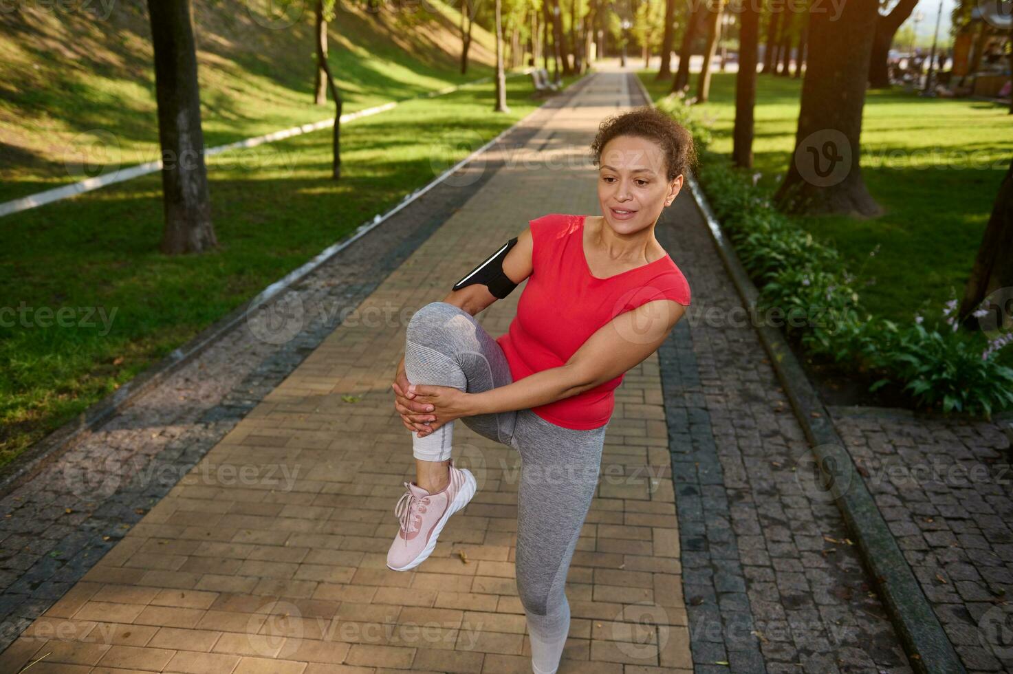 Determined cheerful African American fit woman in gray leggins and red tight t-shirt exercising outdoor in the city park. Fitness, body weight training outdoor on a warm summer day photo