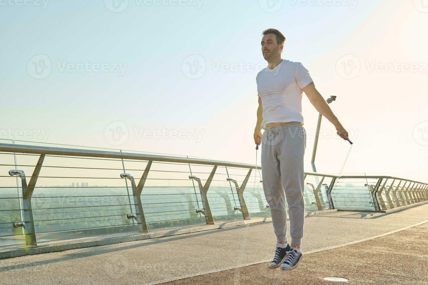 Full length portrait of a muscular build Caucasian man, fit athlete doing jumping exercises, cardio training with a skipping rope on the glass city bridge early in the morning on a summer day photo