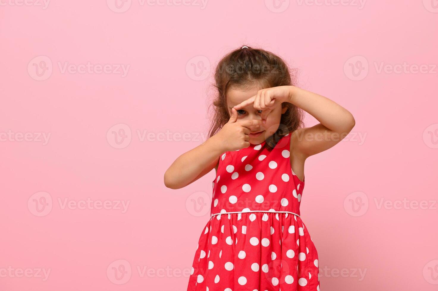 bonito niña mirando mediante el dedo marco, posando a cámara terminado rosado antecedentes con Copiar espacio. internacional muchachas día concepto. facial emociones, positividad, contento infancia. foto