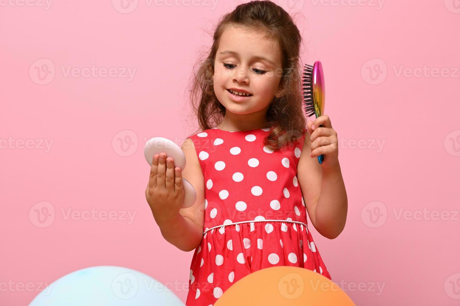 Beautiful Birthday girl in stylish pink dress with polka dots, combs her curly hair, admires her reflection on small pink cosmetic mirror. Portrait isolated over pink background with copy space. photo