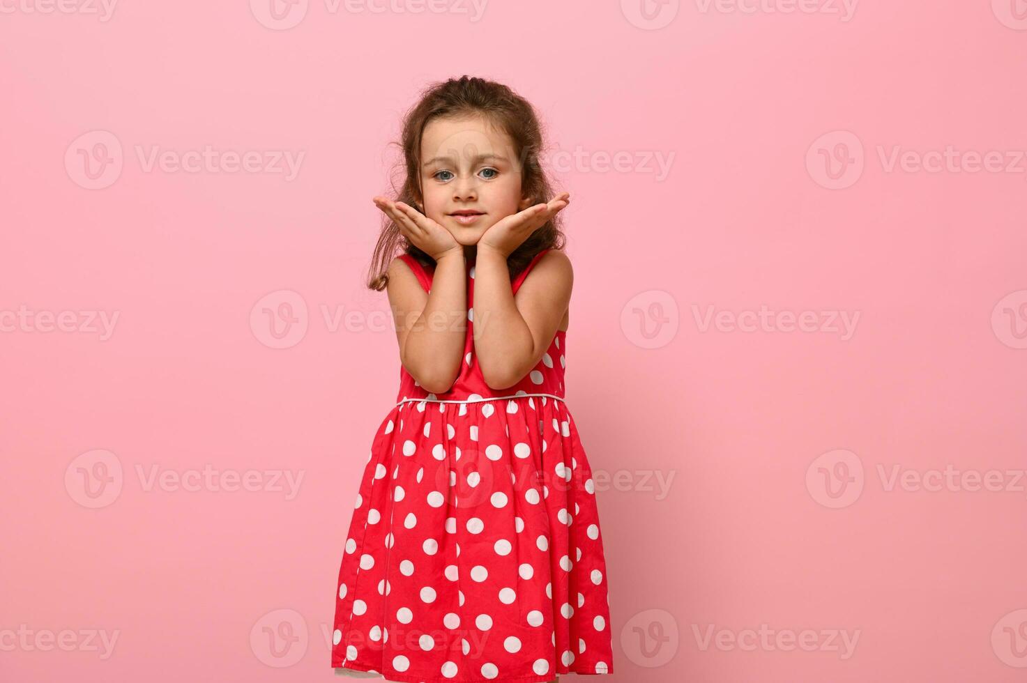 Adorable girl in a pink dress with polka dots poses at the camera, standing against a pink background and holding her chin with her hands. Portrait of a beautiful girl with positive joyful emotions photo