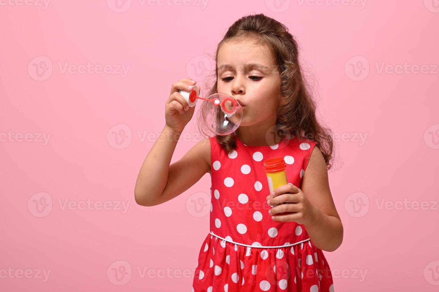 Birthday event concept, Happy Woman's Day, Children protection Day. Adorable child, pretty birthday girl wearing fashionable pink dress with white polka dots starting soap bubbles on pink background photo