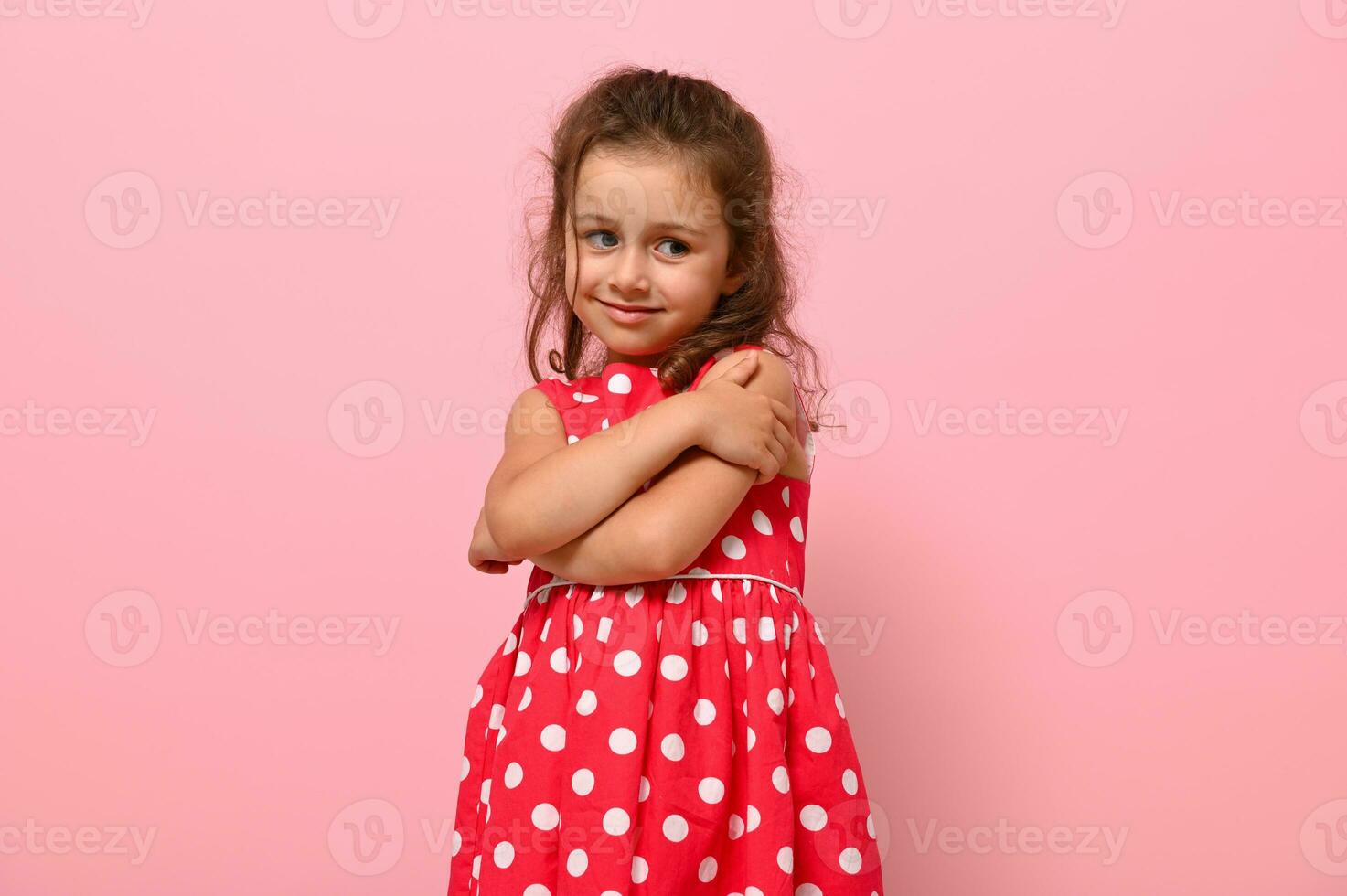 Gorgeous shyly 4 years kid in polka-dots dress hugs herself, sweet smiles posing on pink wall background with space for text. Confident portrait of beautiful baby girl for advertising photo