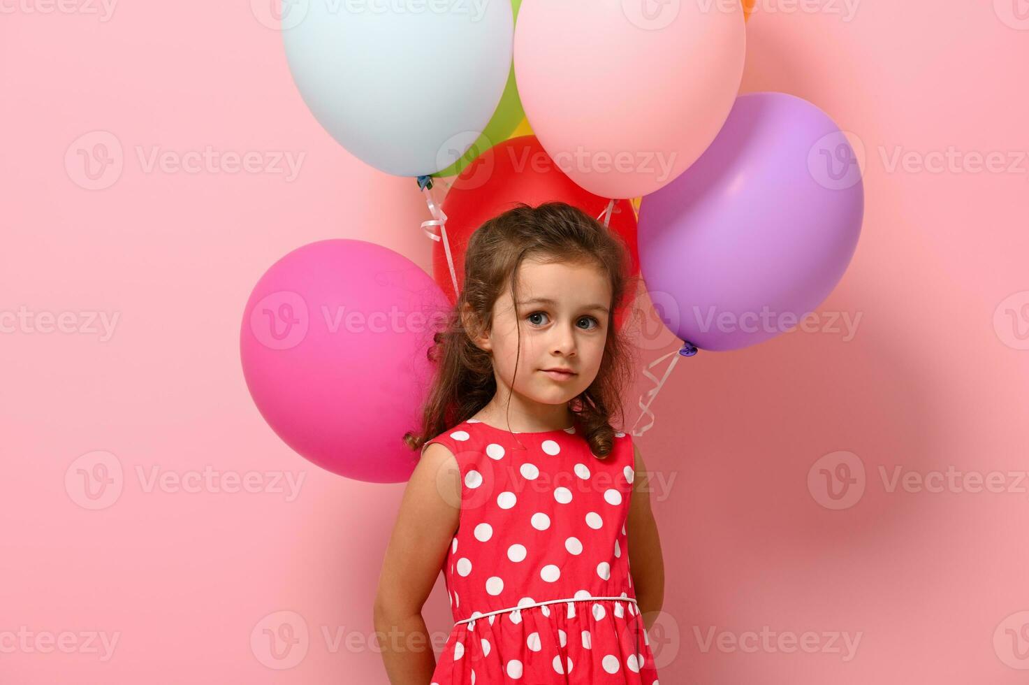 Gorgeous 4 years girl in pink polka dots dress, holding multicolored colorful balloons behind her back, looking at camera, isolated over pink background, copy space. Happy Woman's, Girl's day concept photo