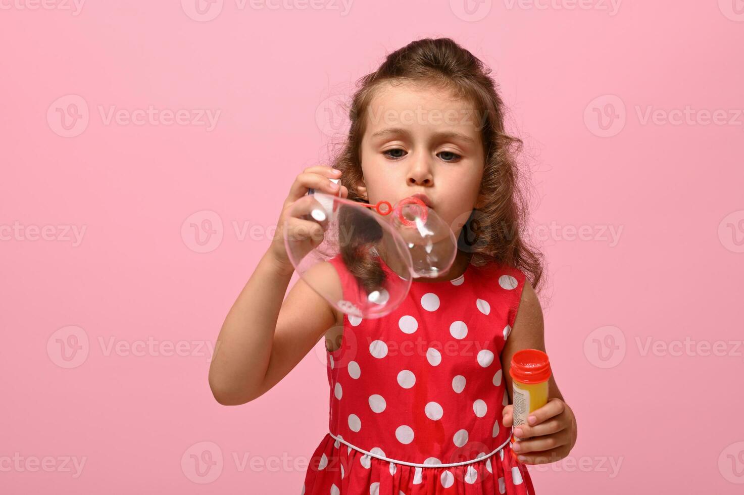 Cheerful happy Birthday girl in pink polka dots dress blowing soap bubbles, isolated over pink background with copy space. Gorgeous child playing with soap bubbles. Summer children leisures concept photo