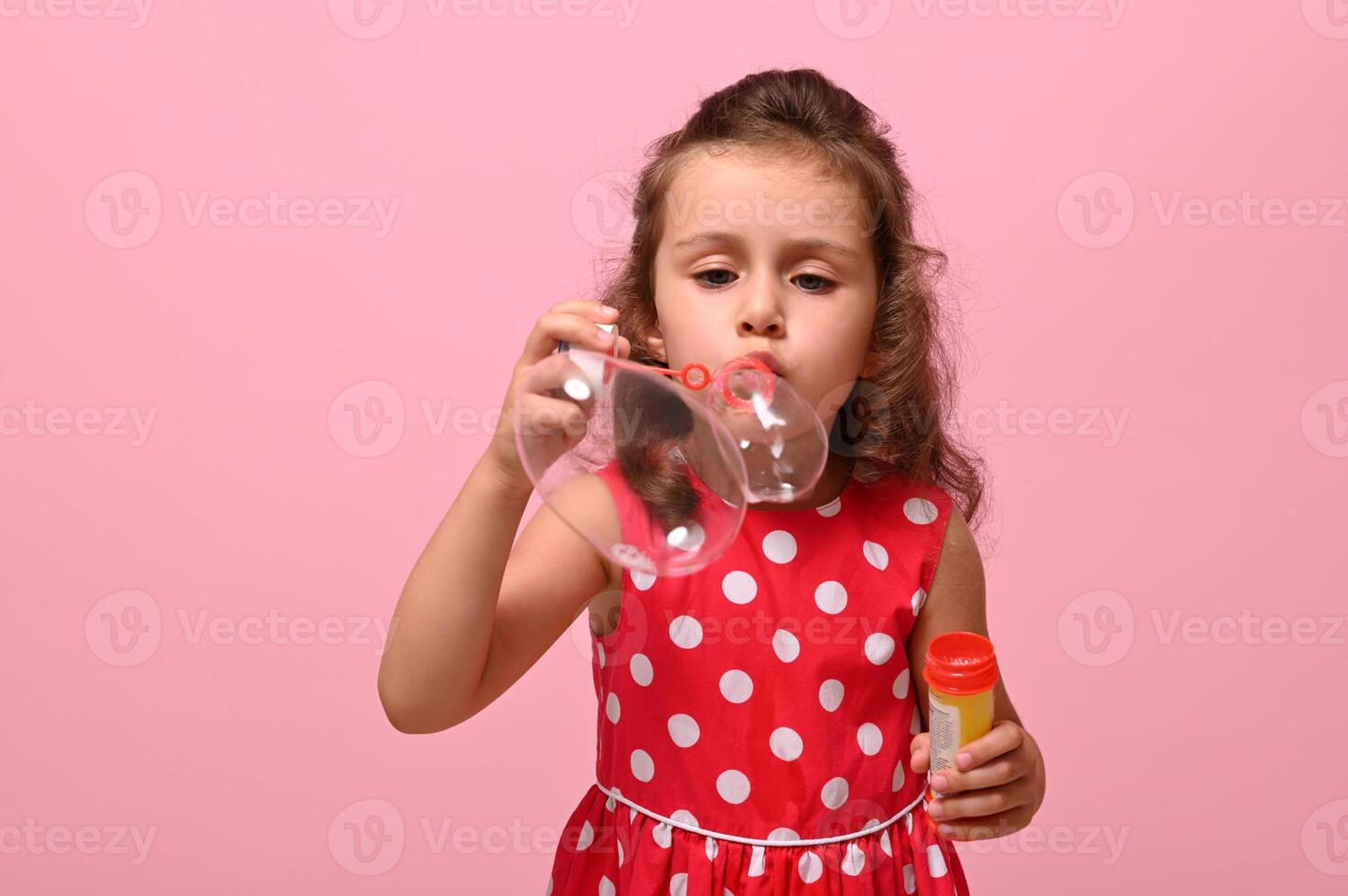 Cheerful happy Birthday girl in pink polka dots dress blowing soap bubbles, isolated over pink background with copy space. Gorgeous child playing with soap bubbles. Summer children leisures concept photo