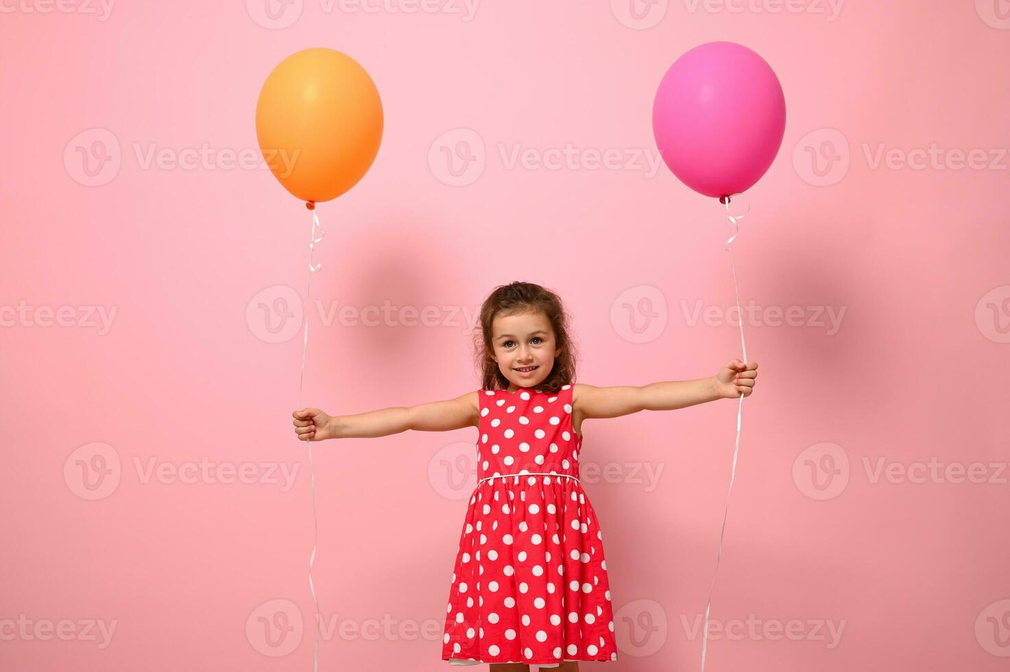 Adorable gorgeous 4 years baby girl in pink dress with white polka dots, holding colorful balloons in her outstretched hands, smiling looking at the camera, isolated on pink background with copy space photo