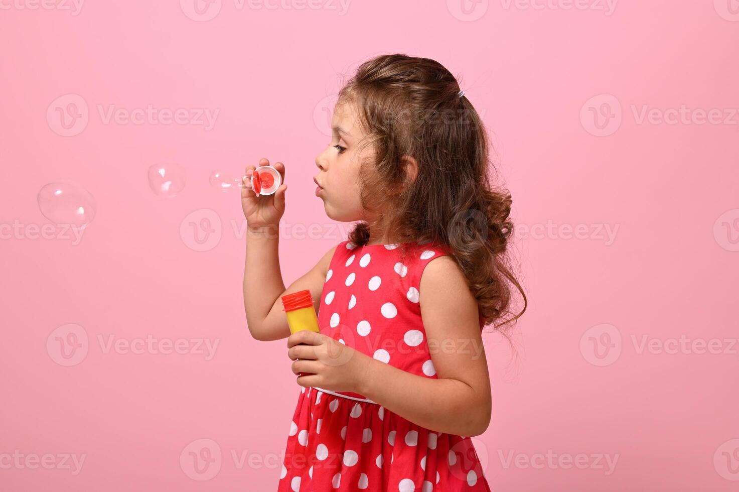 Side portrait of a cheerful beautiful baby girl blowing soap bubbles, isolated over pink background with copy space. Gorgeous child playing with soap bubbles. Summer children leisures concept photo