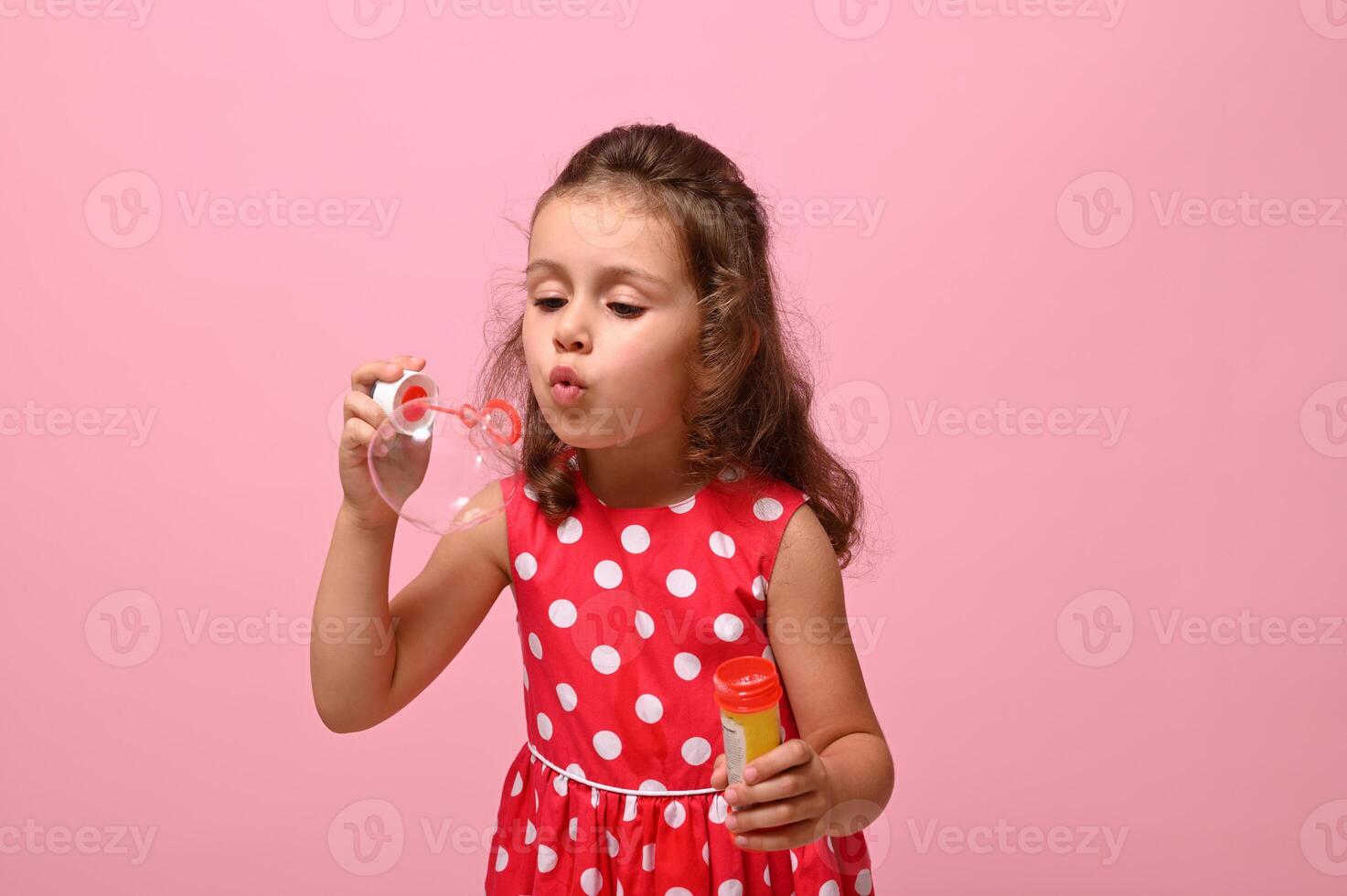 Cheerful Birthday baby girl in stylish pink dress blowing soap bubbles, isolated over pink background with copy space. Gorgeous child playing with soap bubbles. Summer children leisures concept photo