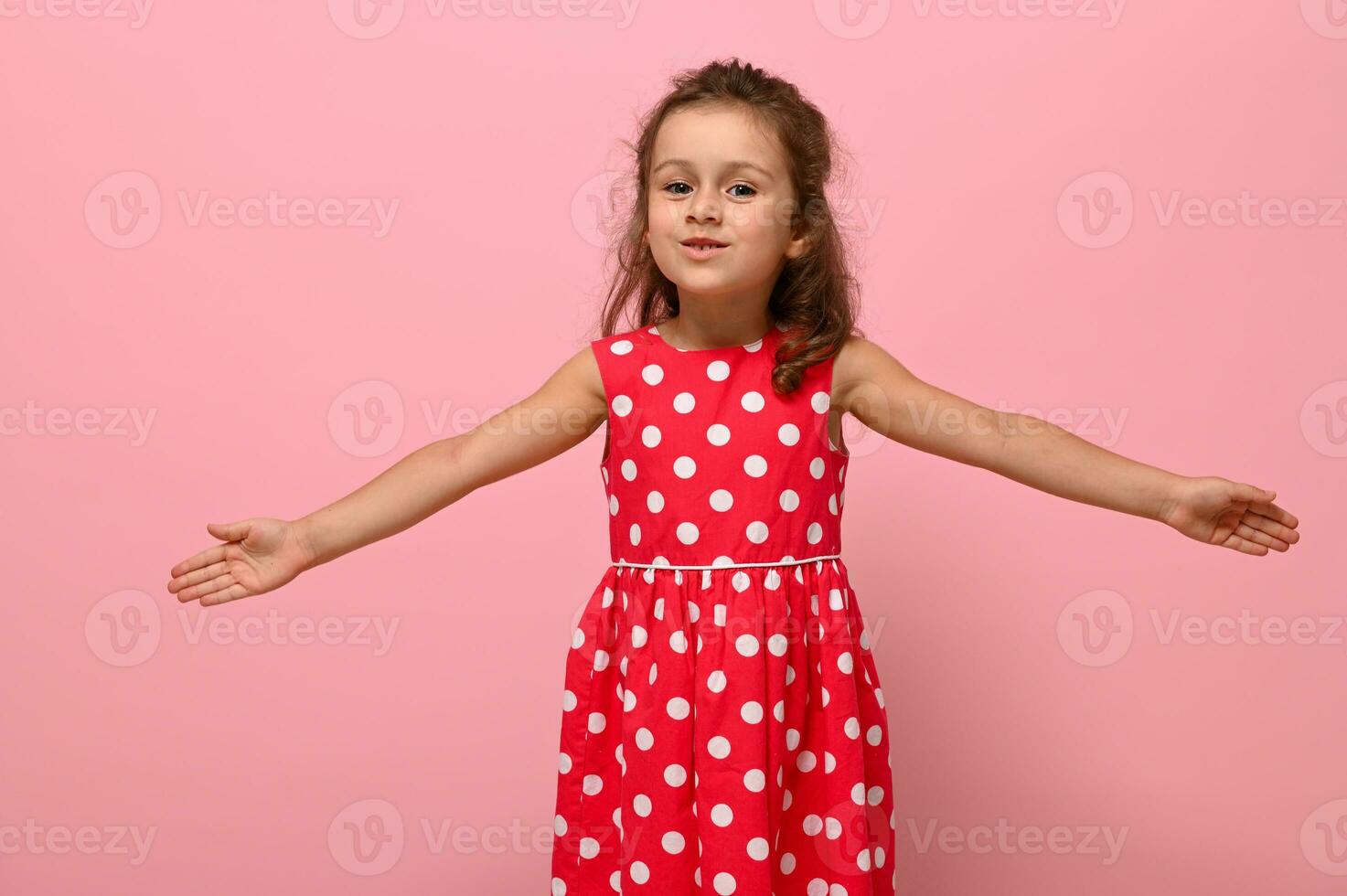 Smiling happy little charming girl with outstretched hands in a pink dress with polka dots, looking at the camera, posing on a pink background. Portrait of good-natured joyful, affable, friendly child photo