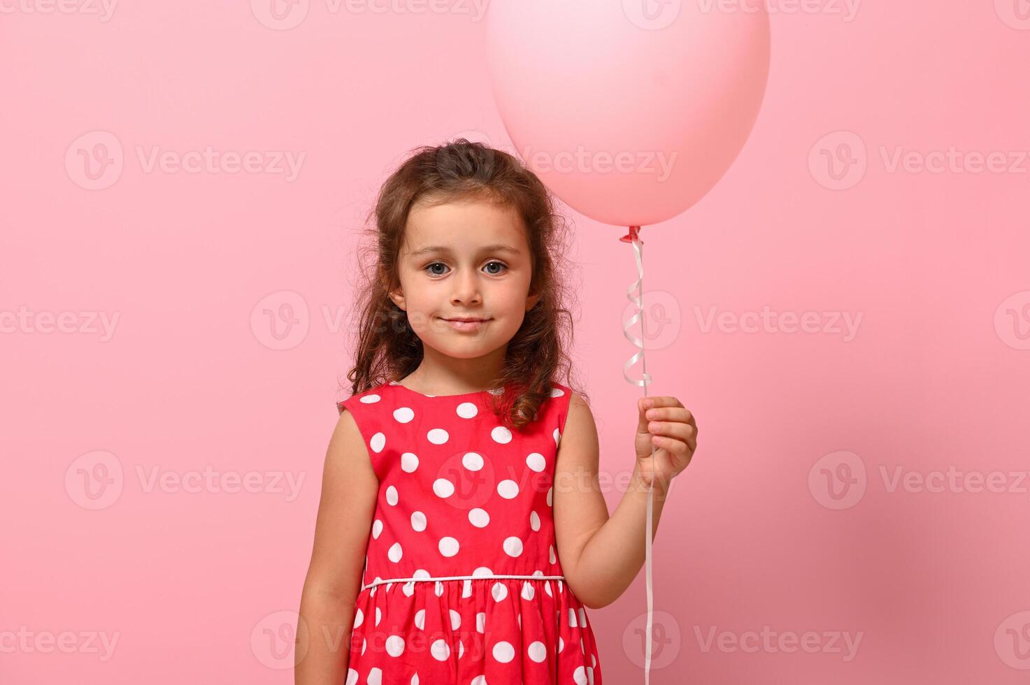 Birthday girl dressed in dress with polka-dots pattern holding pastel pink balloon, smiling, isolated over pink background with copy space. Close-up portrait of beautiful 4 years child for advertising photo