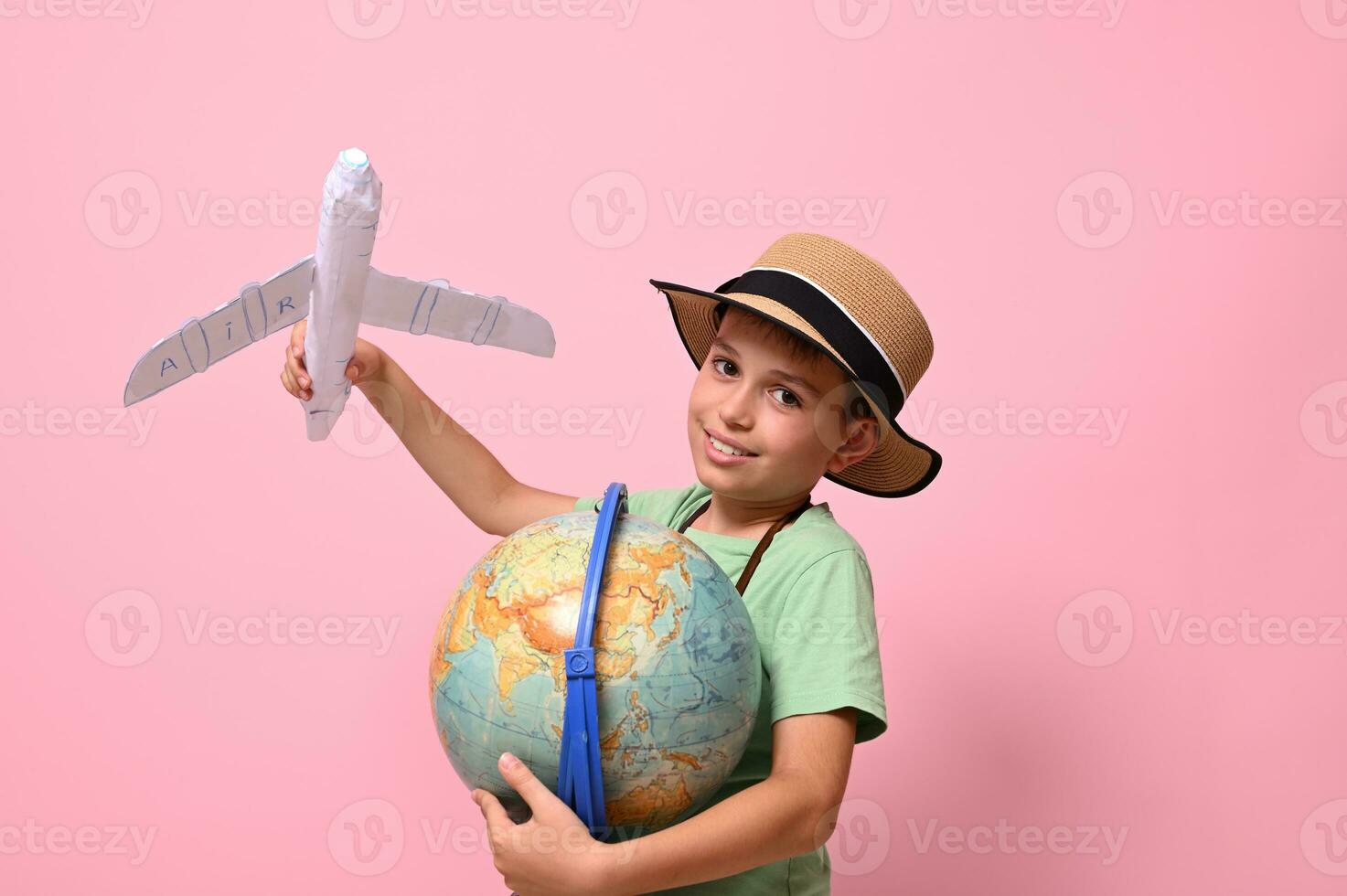 Smiling school boy looking at camera while playing with paper airplane and a globe, standing isolated over pink background with copy space. Tourism, travel, geography knowledge concepts photo