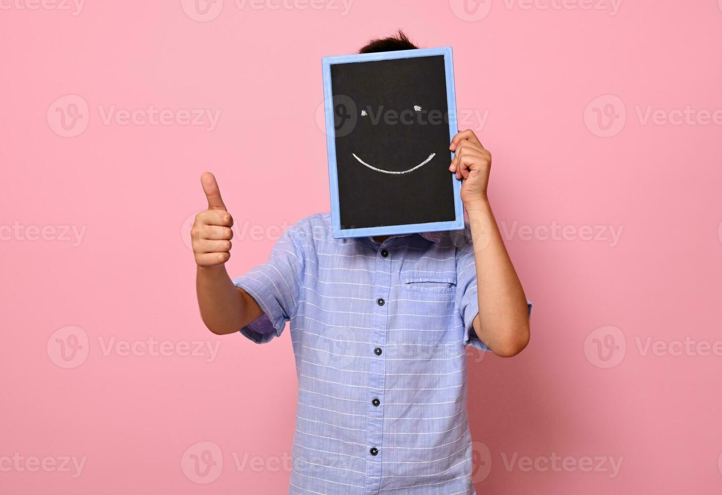 A school boy covers his face with a chalkboard with drawn smiling emoticons, expressing happiness and shows thumb up to the camera. Isolated over pink background with copy space photo