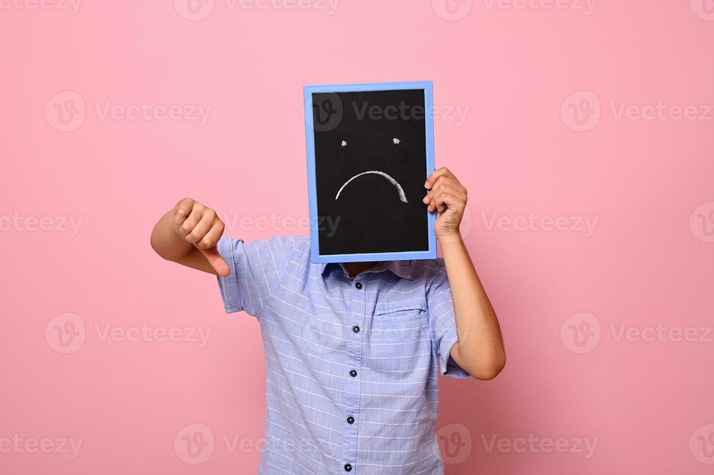 A boy covers his face with a chalkboard with drawn smiling emoticons, expressing sadness and shows thumb down to the camera. Isolated over pink background with copy space photo