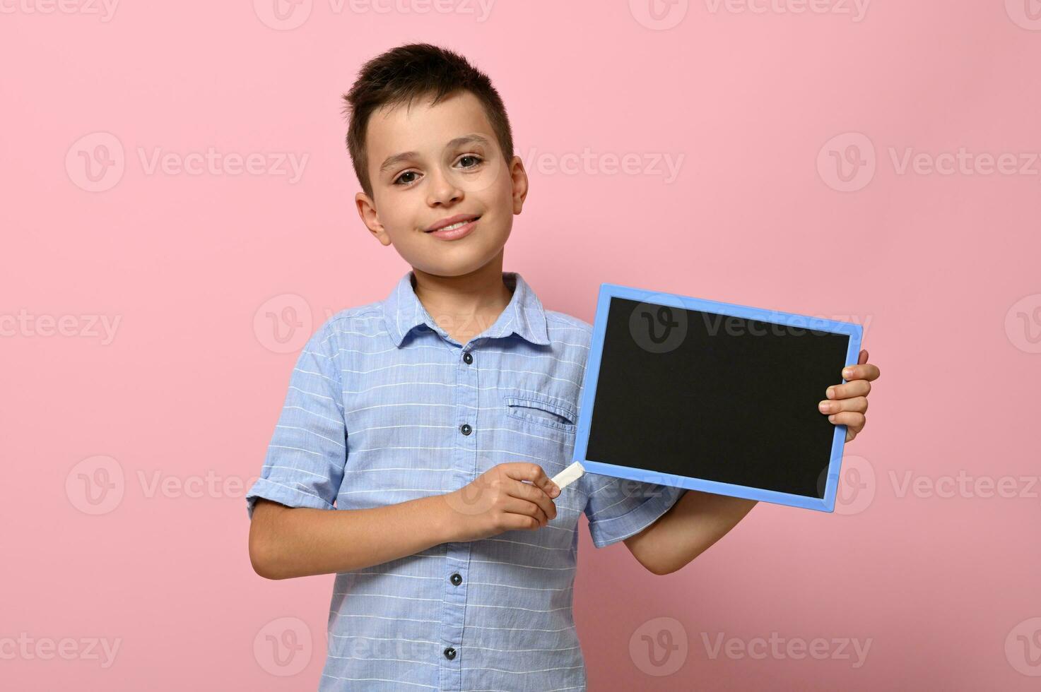 Adorable smiling boy pointing on an empty blank board with a chalk. Space for text on a chalkboard. Pink background with copy space. Concepts of back to school photo