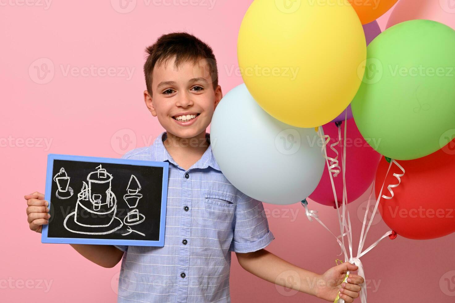 Cute boy holds multicolored colorful balloons in one hand and a chalkboard with drawn birthday cake in the other. Isolated over pink background with copy space photo