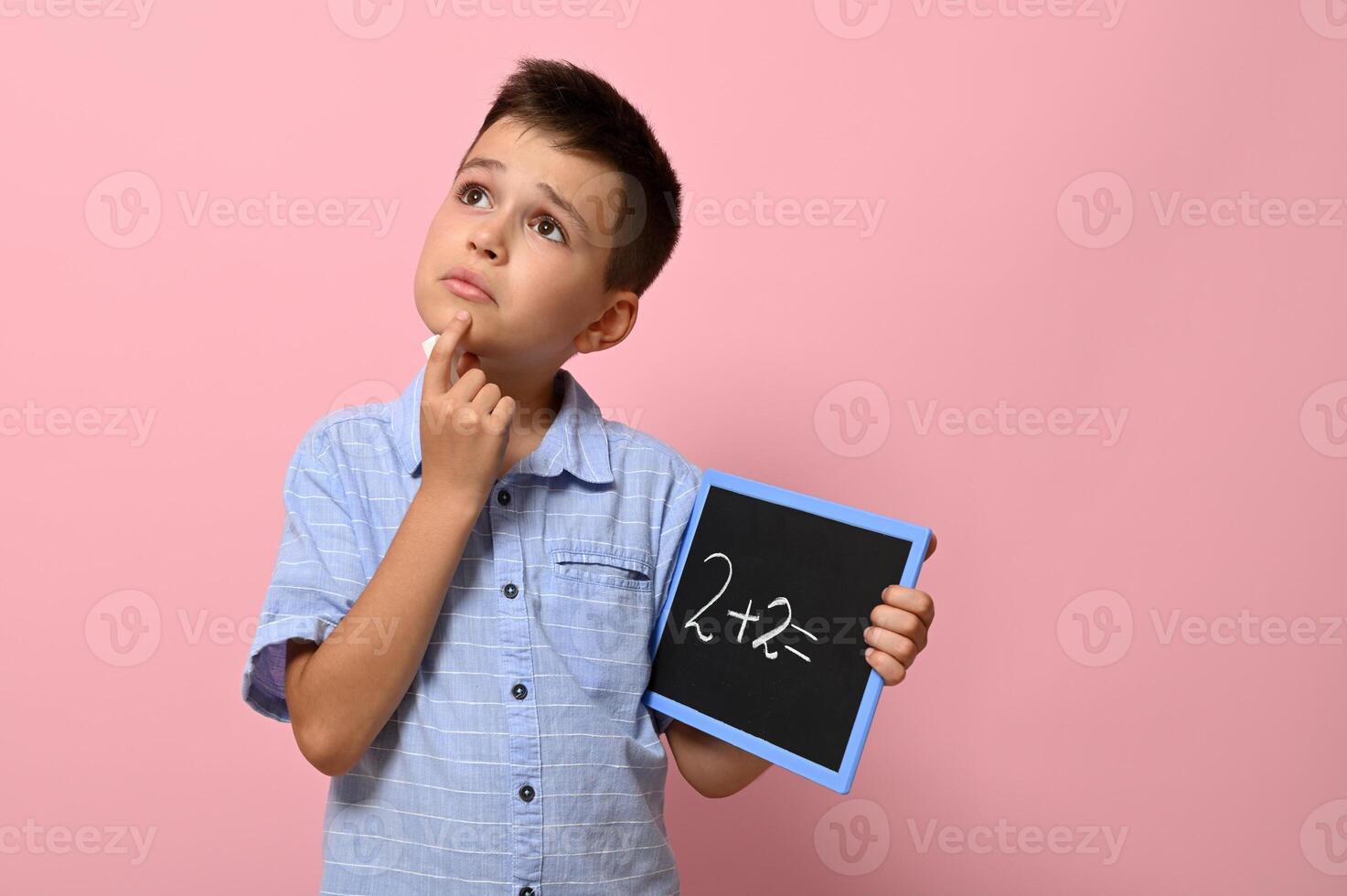 Cute schoolboy holding a chalkboard with chalk writing on it, thoughtfully solves a math problem. Isolated on pink background with copy space photo