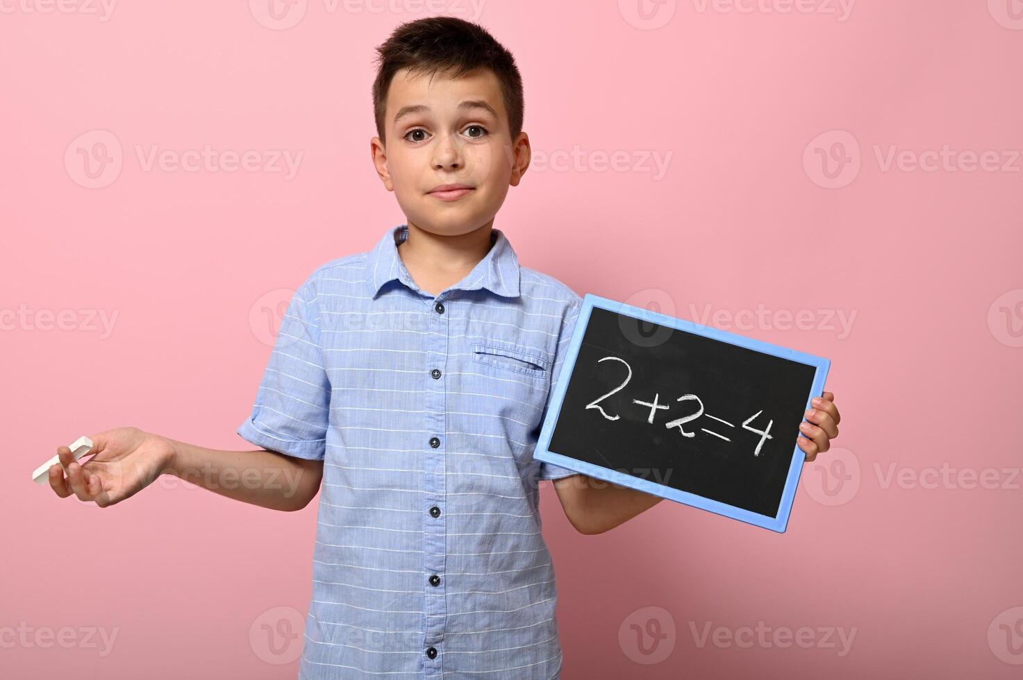 Adorable boy, elementary school student holds chalk and blackboard and solves math problem. Pink background space for text. Back to school. Concepts photo