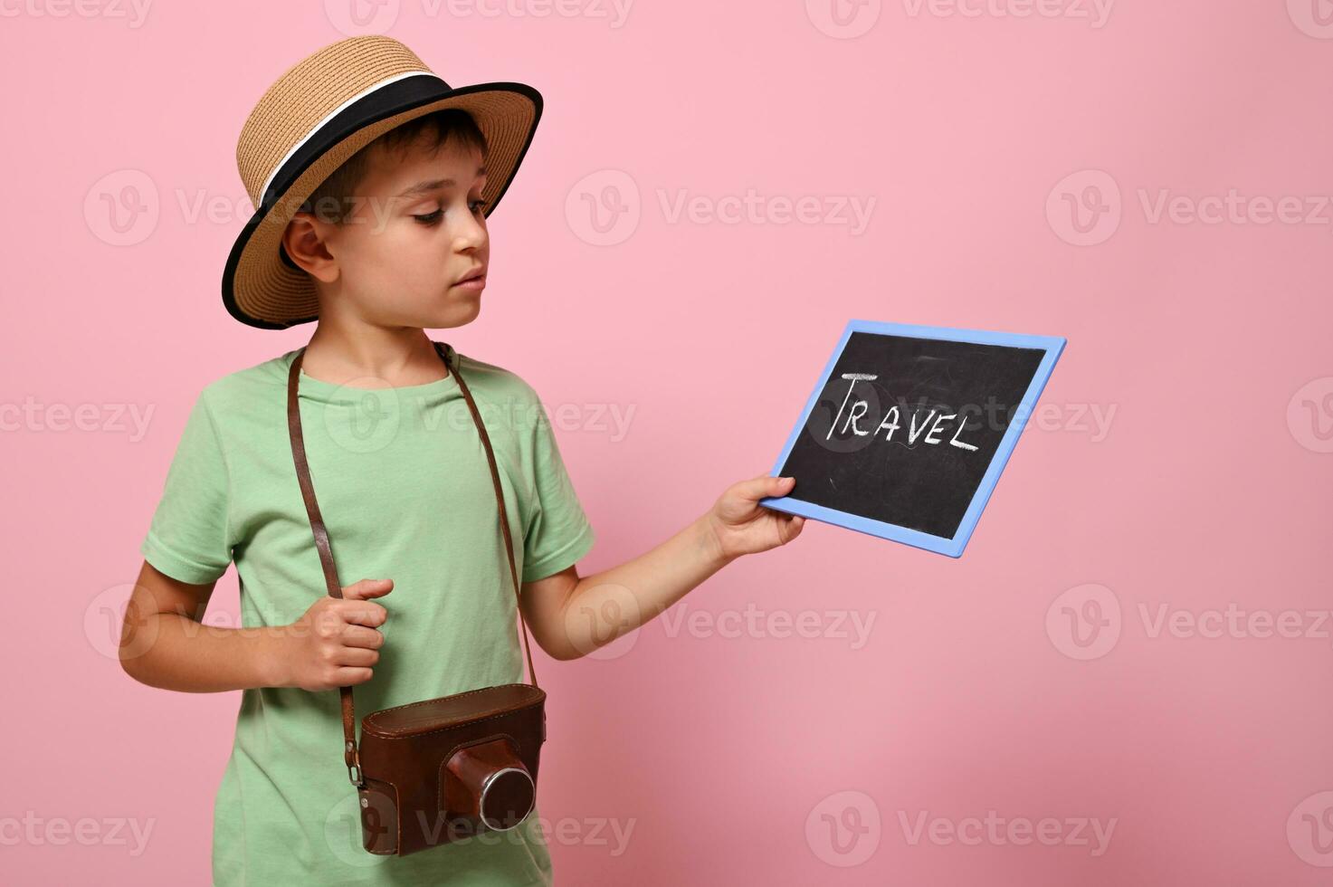 hermoso chico en verano sombrero con un Clásico cámara alrededor el cuello mirando a pizarra con letras viaje . turismo y verano conceptos en un rosado antecedentes con Copiar espacio foto