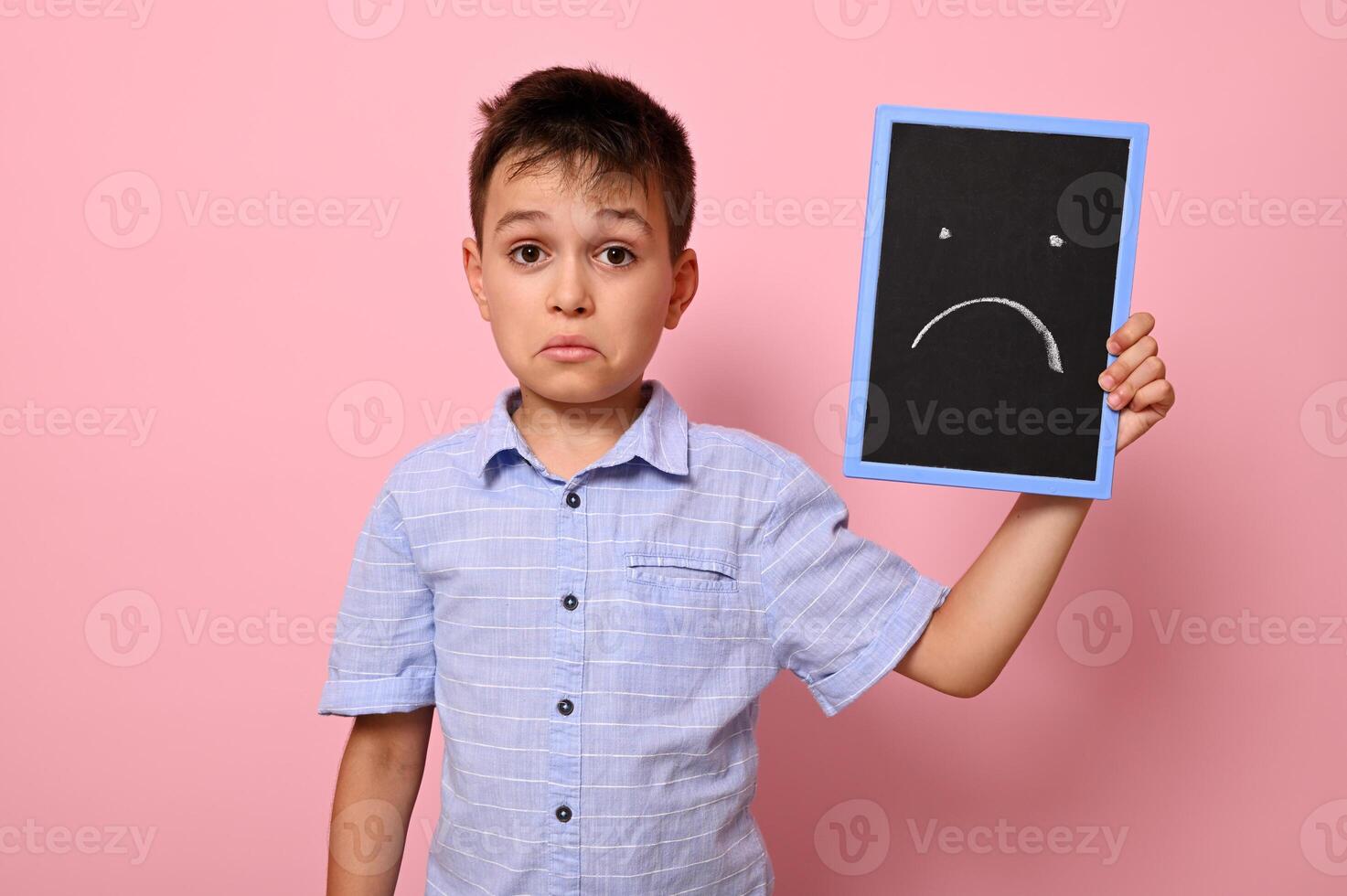 A frustrated boy in a blue shirt holds a blackboard near his face with a painted drawn expressing frustration, sadness and disappointment. Pink background, copy space photo