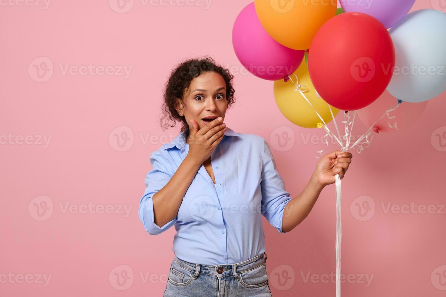 Surprised beautiful stunning mixed race woman of African ethnicity with colorful air balls in hand covers her mouth, expressing surprise and astonishment. Isolated on pink background with copy space photo