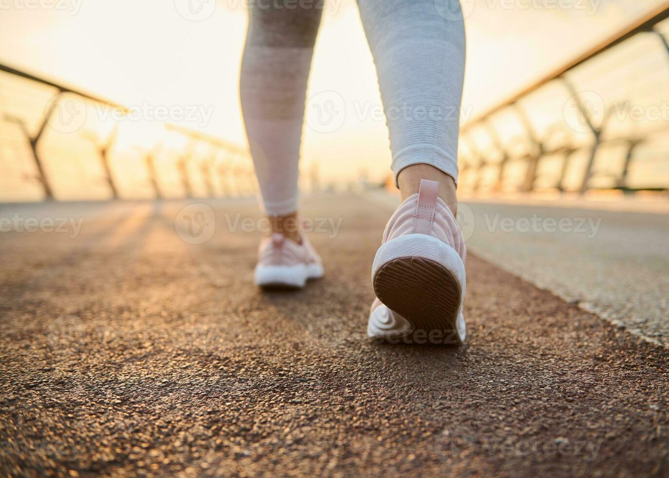 Close-up female athlete feet in pink sneakers on a treadmill at sunrise background. Cropped image of the legs of a sportswoman running jogging on a city bridge with sun beams falling on treadmill photo