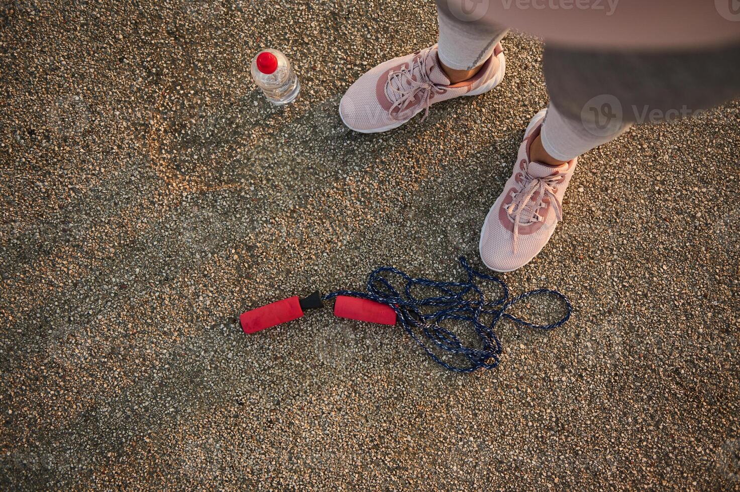 High angle view of female athlete legs wearing pink sneakers and standing on an asphalt treadmill next to a lying down skipping rope and water bottle photo