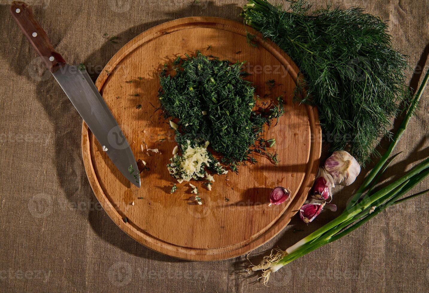 Overhead view of a bunch of fresh moist dill on a linen tablecloth and garlic next to a wooden cutting board with a kitchen knife and chopped garlic and dill. Seasonal greens food background. Flat lay photo