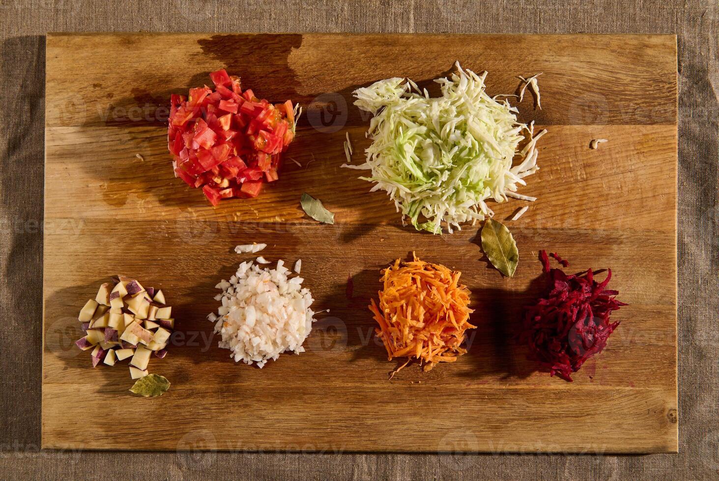 Flat lay of chopped ingredients for a traditional Ukrainian soup- borscht. Chopped and grated raw fresh vegetables -beet, carrot onions, tomatoes, potatoes, and cabbage on a wooden cutting board. photo