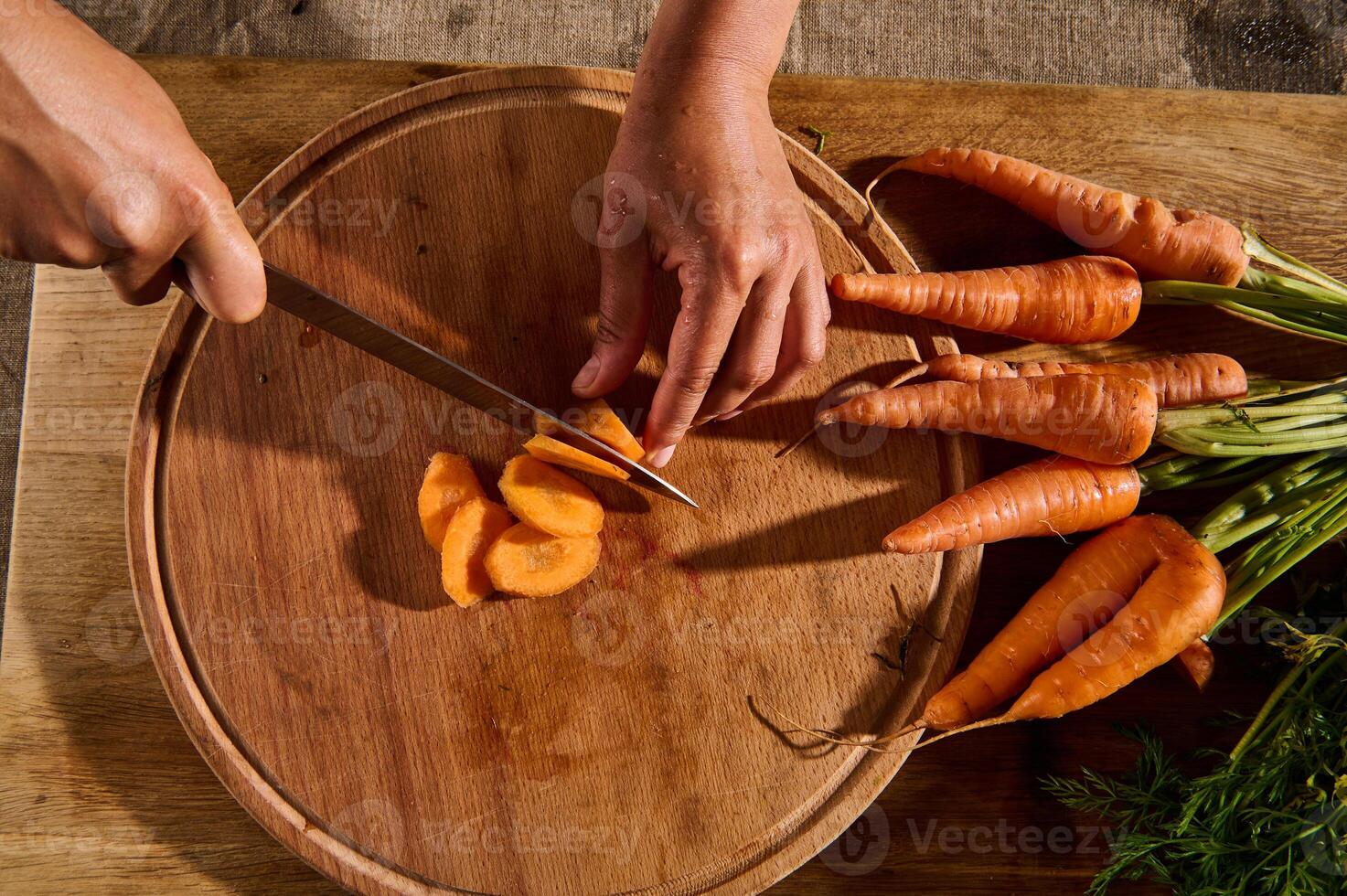 Overhead view of hands chopping, slicing fresh raw baby carrot on wooden board. Close-up of hand with knife cutting vegetable. photo