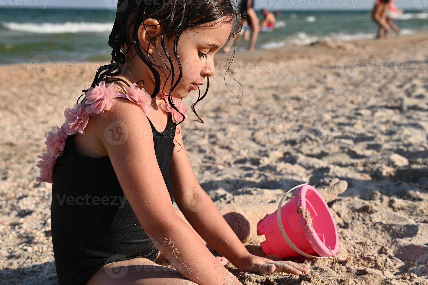 preescolar niña jugando con rastrillo y rosado juguete Cubeta con arena, edificio arenoso formas y castillos, disfrutando broncearse y verano vacaciones contento infancia, bienestar y recreación conceptos foto