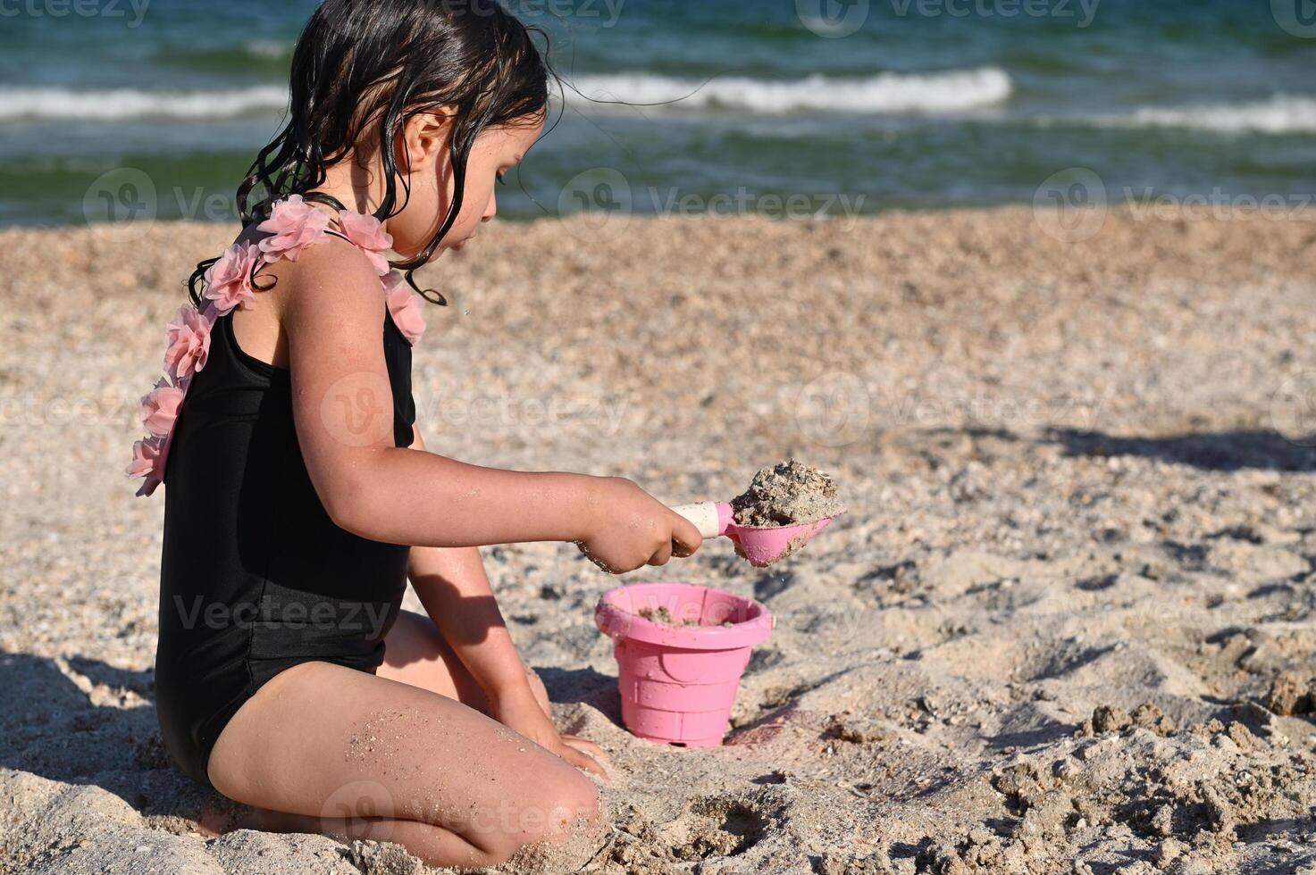 Side portrait of preschooler girl holding a rake and filling the pink toy bucket with sand, building sandy shapes and castles, enjoying sunbathing and summer vacations. photo