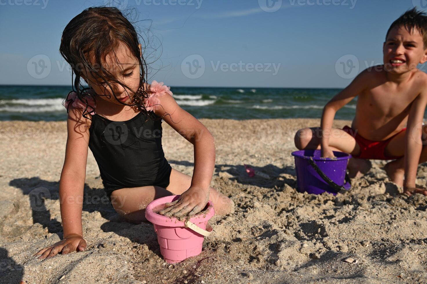 linda niña llena un rosado el plastico juguete Cubeta con arena a crear estatuillas y construye un arena castillo mientras jugando junto a su hermano en contra el fondo de el mar. foto