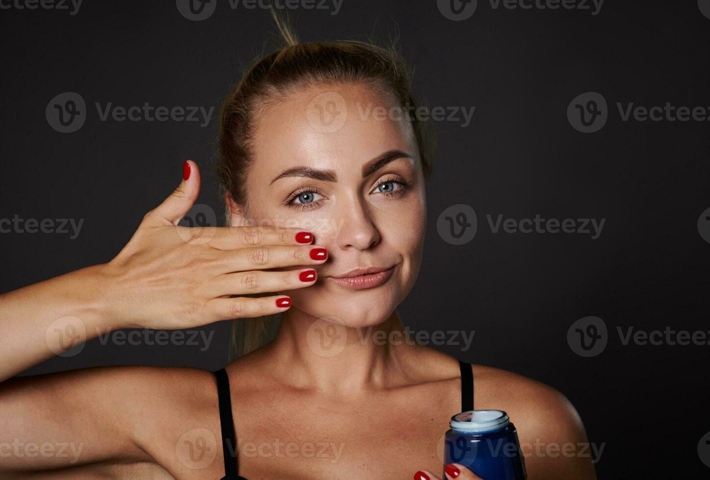 Close-up beauty portrait beautiful Caucasian woman applying moisturizer on face, holding jar of daily smoothing cream or beauty serum, isolated on black background. Skin care, cosmetology, spa concept photo